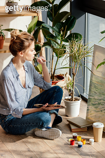 side view of young female with paintbrush and palette sitting at floor ...
