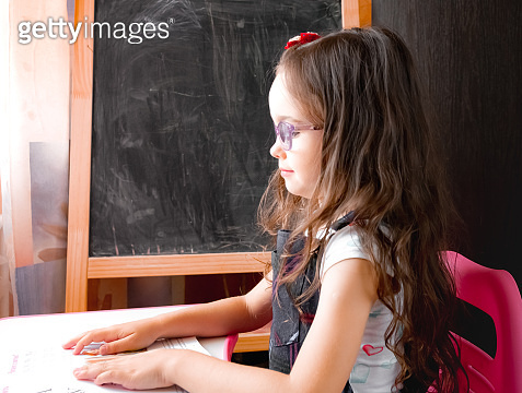 Smart little girl in eyeglasses is sitting at a desk and reading book ...