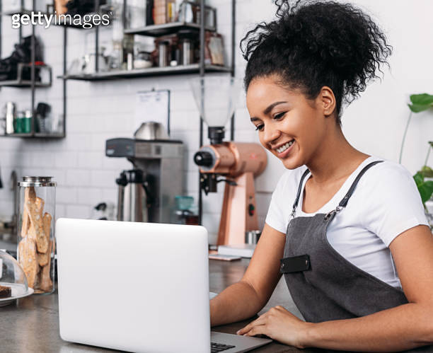 Smiling woman with curly hair wearing an apron typing on a laptop in a ...
