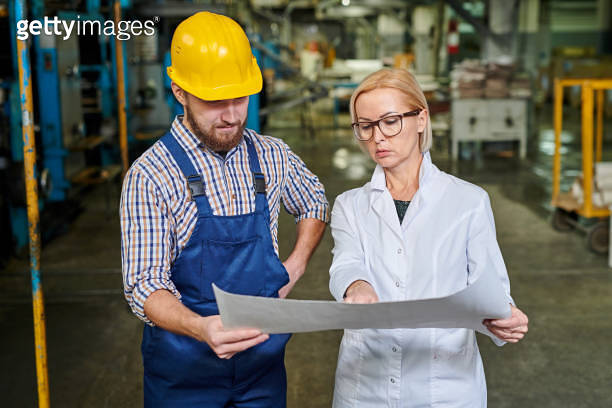 Serious strict lady in lab coat pointing at blueprint while explaining ...
