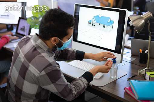 Mixed race man wearing face mask sanitizing hands at desk using ...
