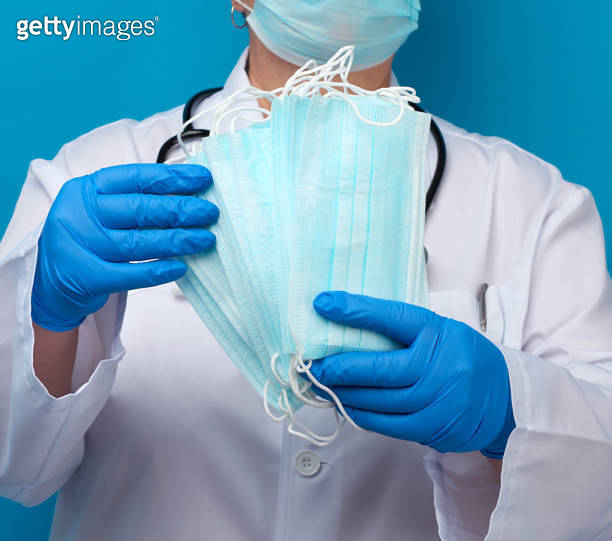 doctor in a white coat and mask holds a stack of protective disposable