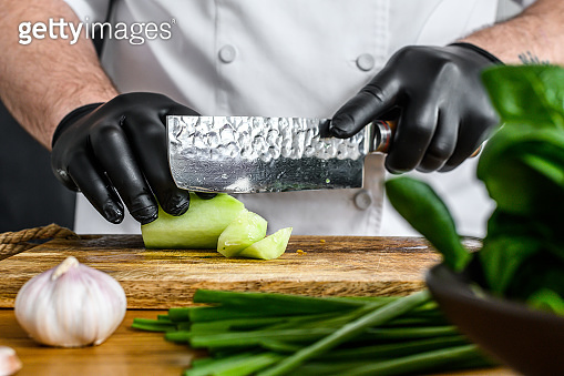 A chef in black gloves is slicing a fresh green cucumber on a wooden ...