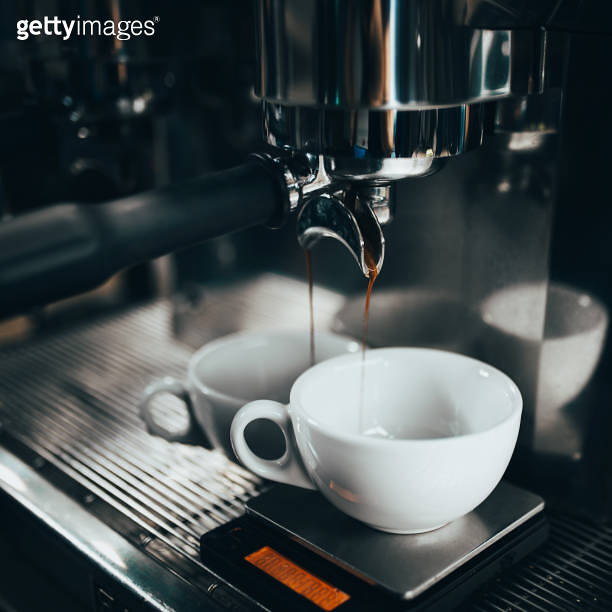 Espresso drips from a coffee machine into a white cup in a thin stream