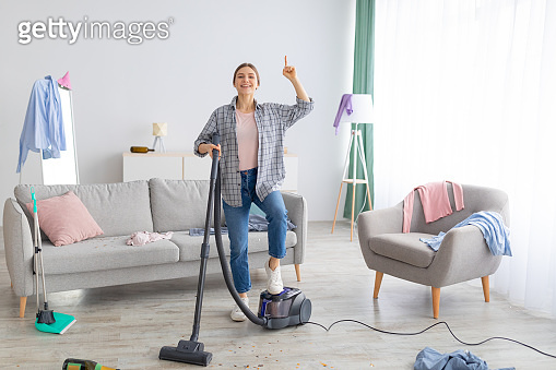 Happy young lady with vacuum cleaner having cleanup idea at messy room ...