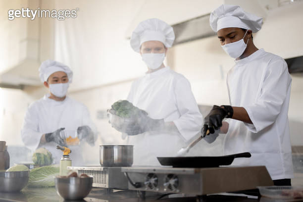 Multiracial team of cooks in uniform and face masks cooking meals for a ...