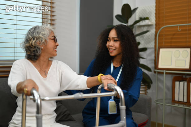 Positive female caregiver teaching senior woman to walk with walker ...