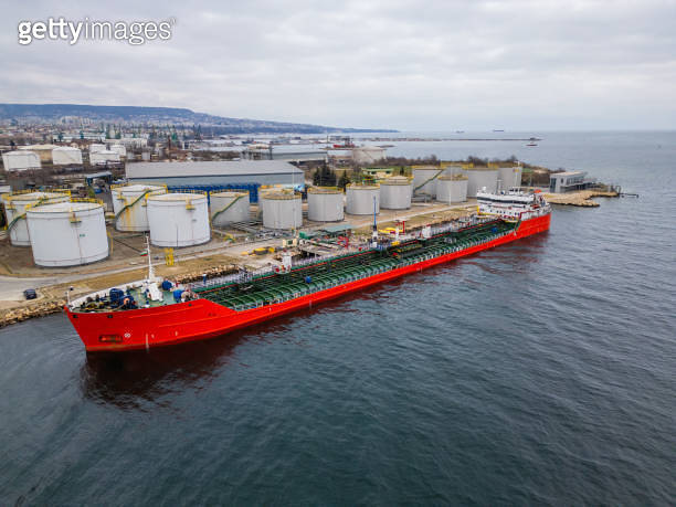 A red tanker ship docked at an industrial port with large storage tanks ...